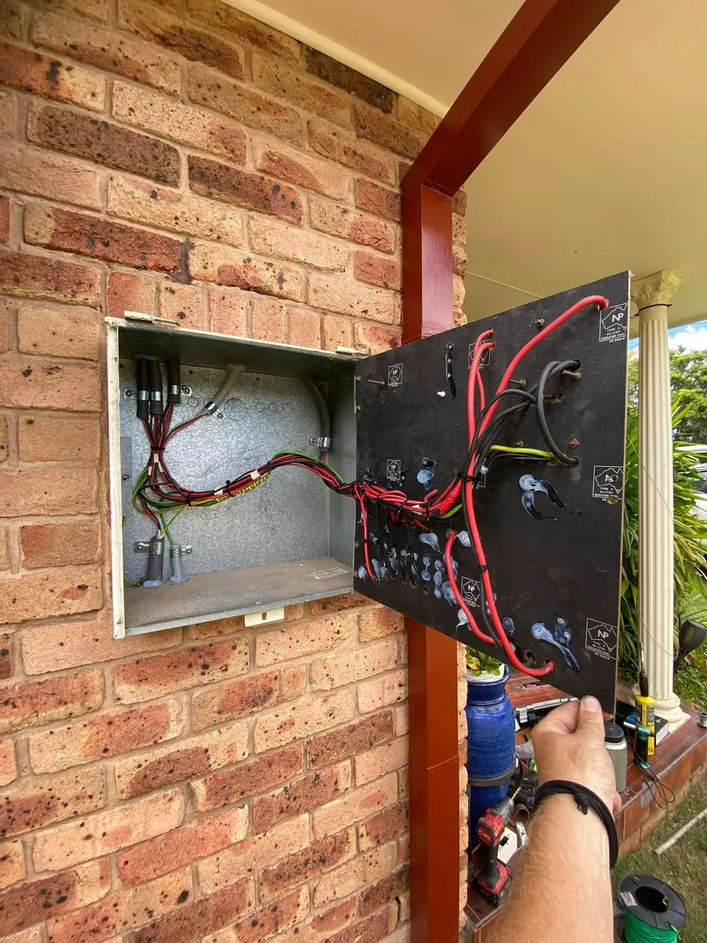 An electrician wiring an open switchboard enclosure on the wall of a brick home